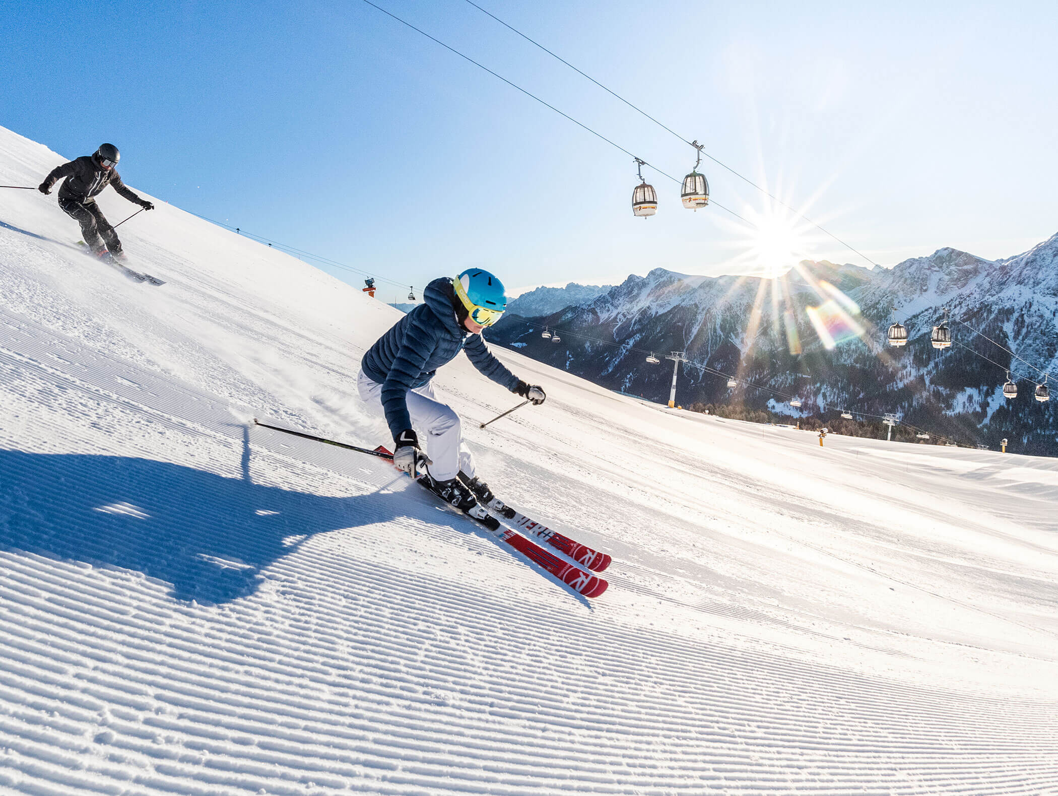Zwei Skifahrer rasen einen sonnigen, schneebedeckten Berghang hinunter, mit Skiliften und Bergen im Hintergrund. - Hotel B&B MyLiving