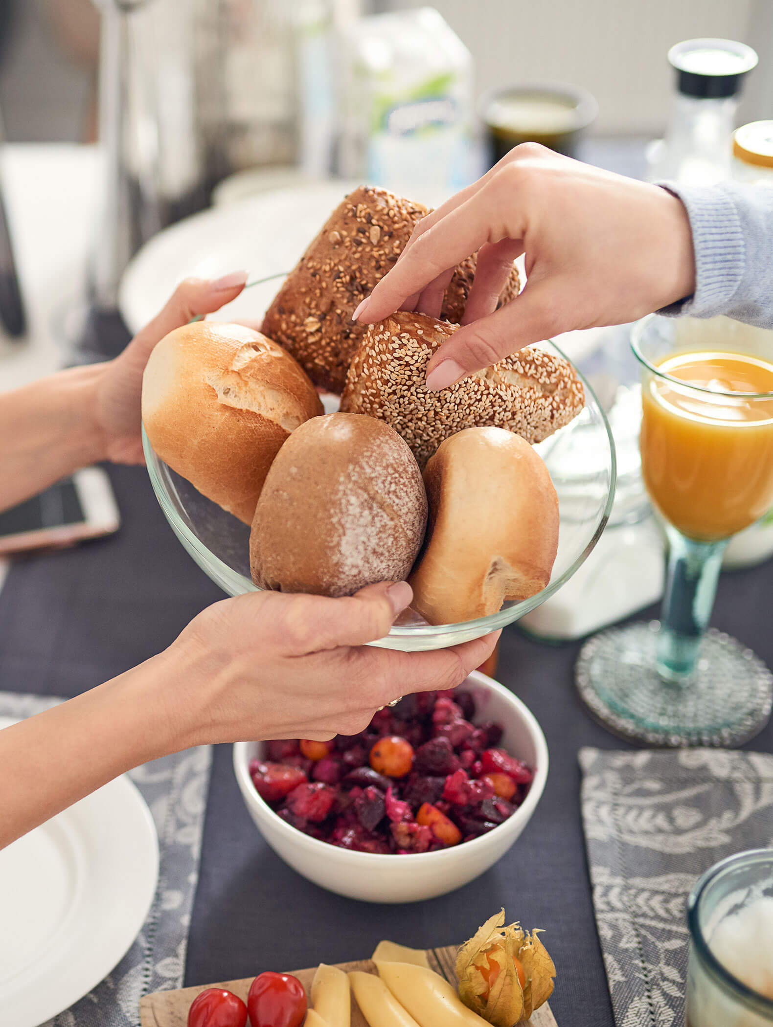 Zwei Hände halten eine Schüssel mit verschiedenen Brötchen über einen Frühstückstisch mit Saft und Obst. - Hotel B&B MyLiving
