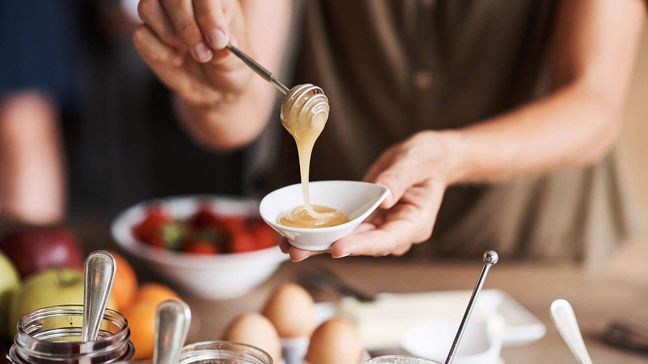 Eine Person träufelt Honig aus einer Schöpfkelle in eine kleine weiße Schale, während im Hintergrund Lebensmittel und Früchte zu sehen sind. - Hotel B&B MyLiving