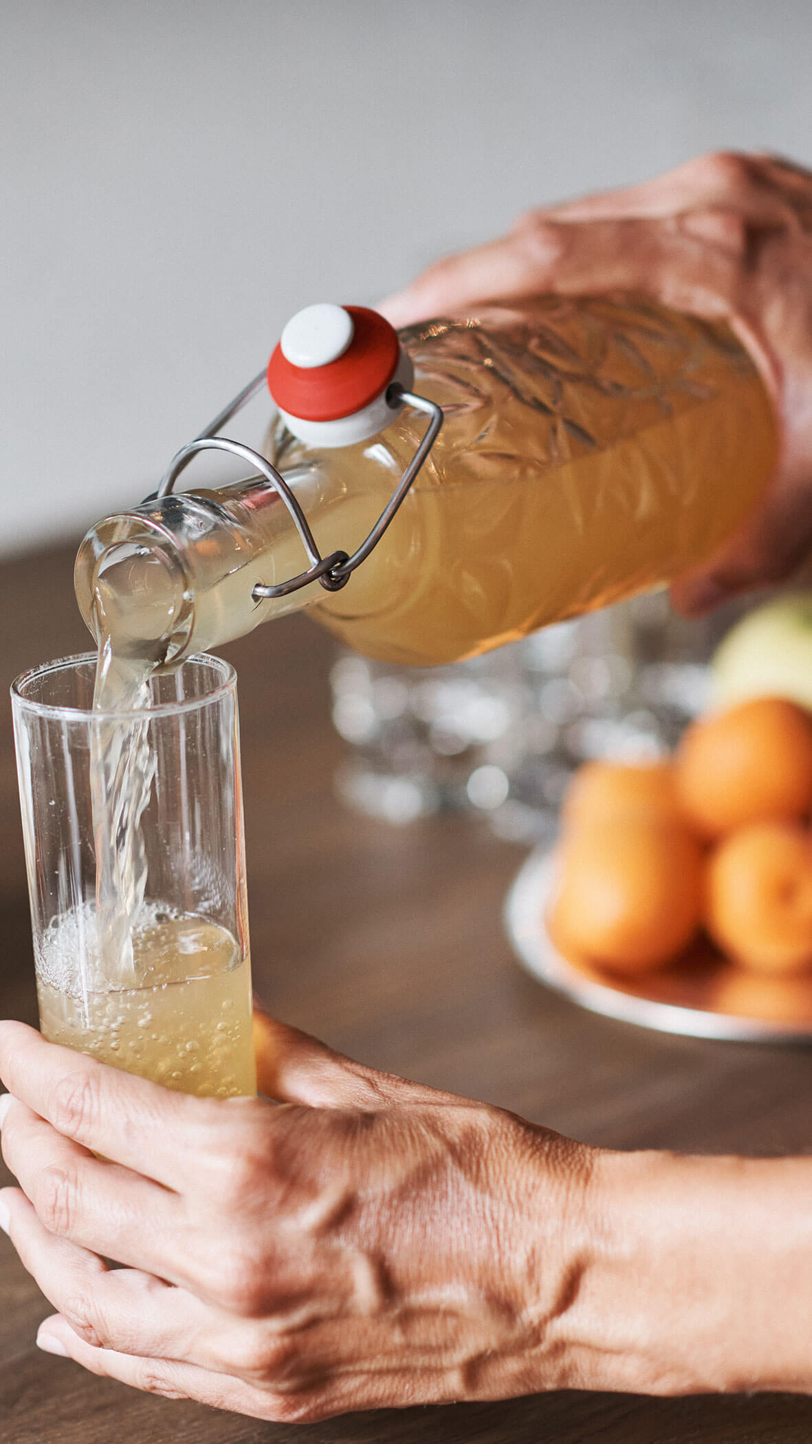 Eine Person gießt ein helles Getränk aus einer Glasflasche in ein hohes Glas neben einem Teller mit Aprikosen. - Hotel B&B MyLiving