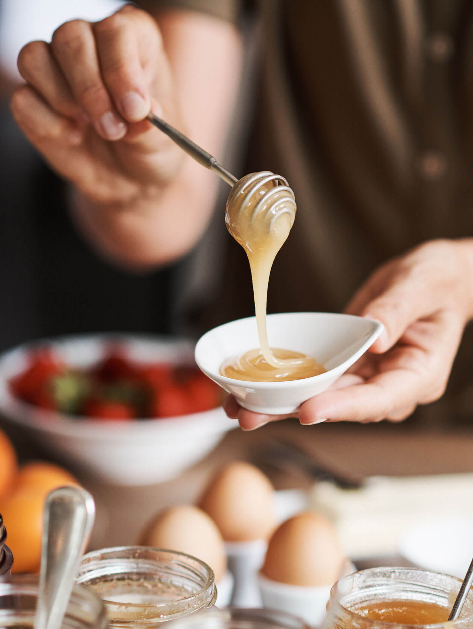Eine Person träufelt Honig aus einer Schöpfkelle in eine kleine weiße Schale, im Hintergrund sind Eier und Früchte zu sehen. - Hotel B&B MyLiving