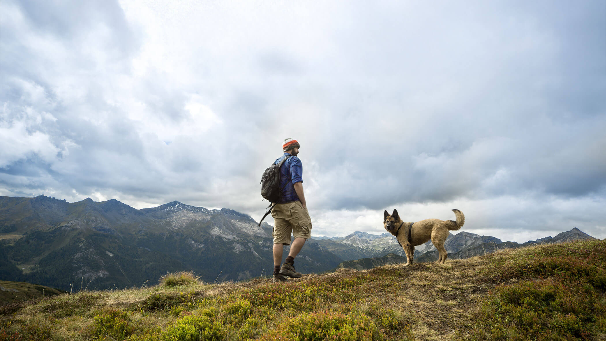 Person, die auf einem grasbewachsenen Bergpfad mit einem Hund wandert, bewölkter Himmel und Berge im Hintergrund. - Hotel B&B MyLiving