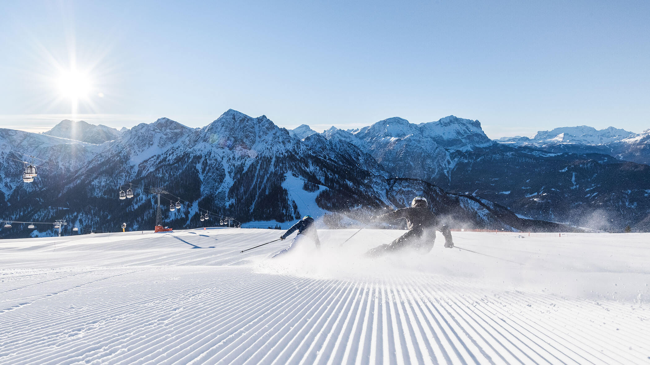 Zwei Personen fahren auf einer frisch präparierten Piste mit verschneiten Bergen und einer strahlenden Sonne im Hintergrund. - Hotel B&B MyLiving