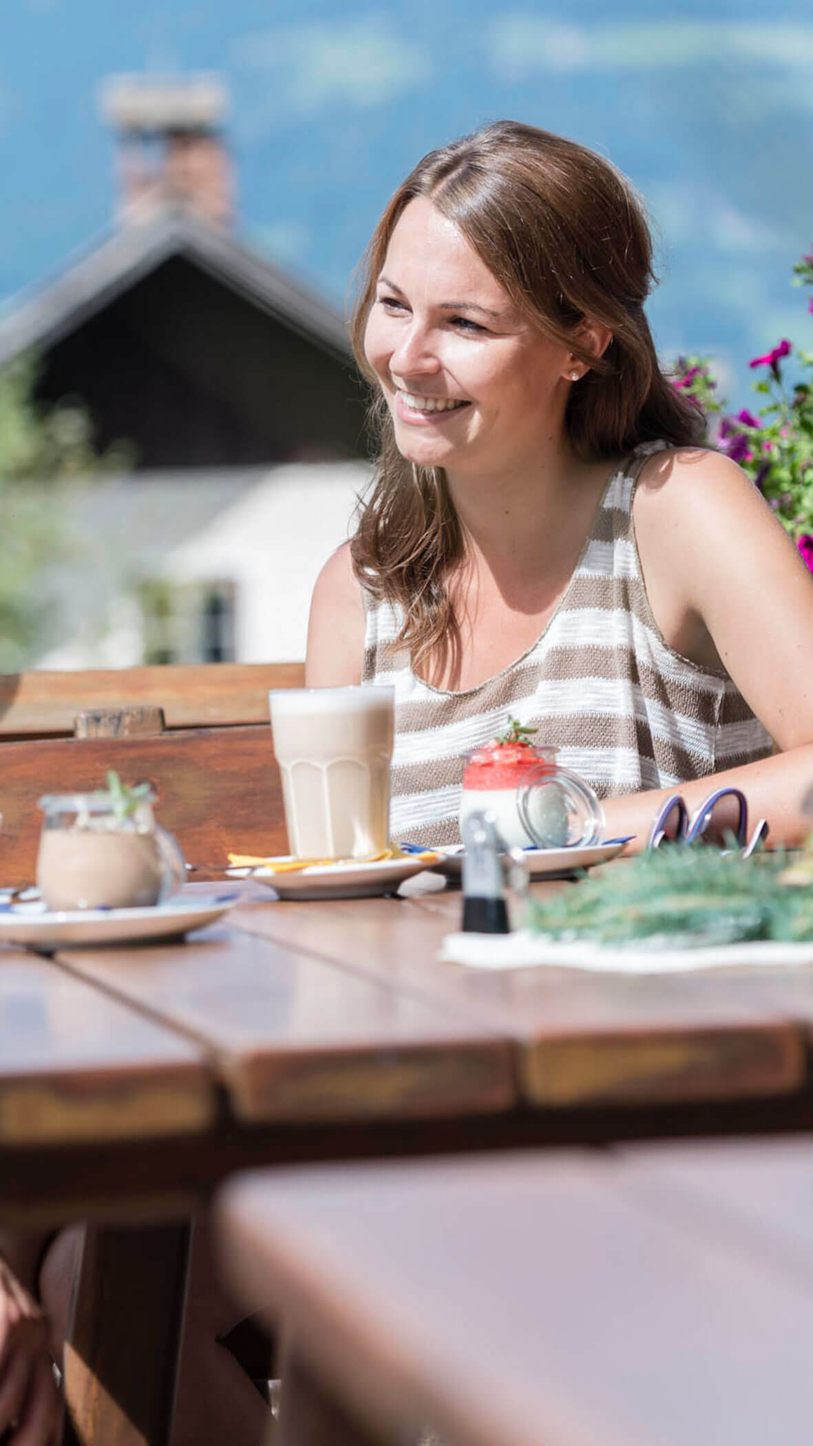 Eine lächelnde Frau sitzt an einem sonnigen Tag mit Getränken und Blumen an einem Cafétisch im Freien. - Hotel B&B MyLiving