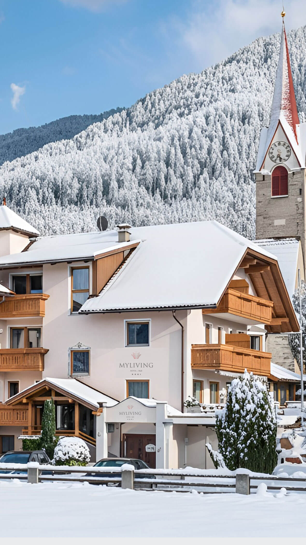 Schneebedeckte Alpenhäuser mit Holzbalkonen, eine Kirche mit Uhrenturm und eine bewaldete Bergkulisse. - Hotel B&B MyLiving