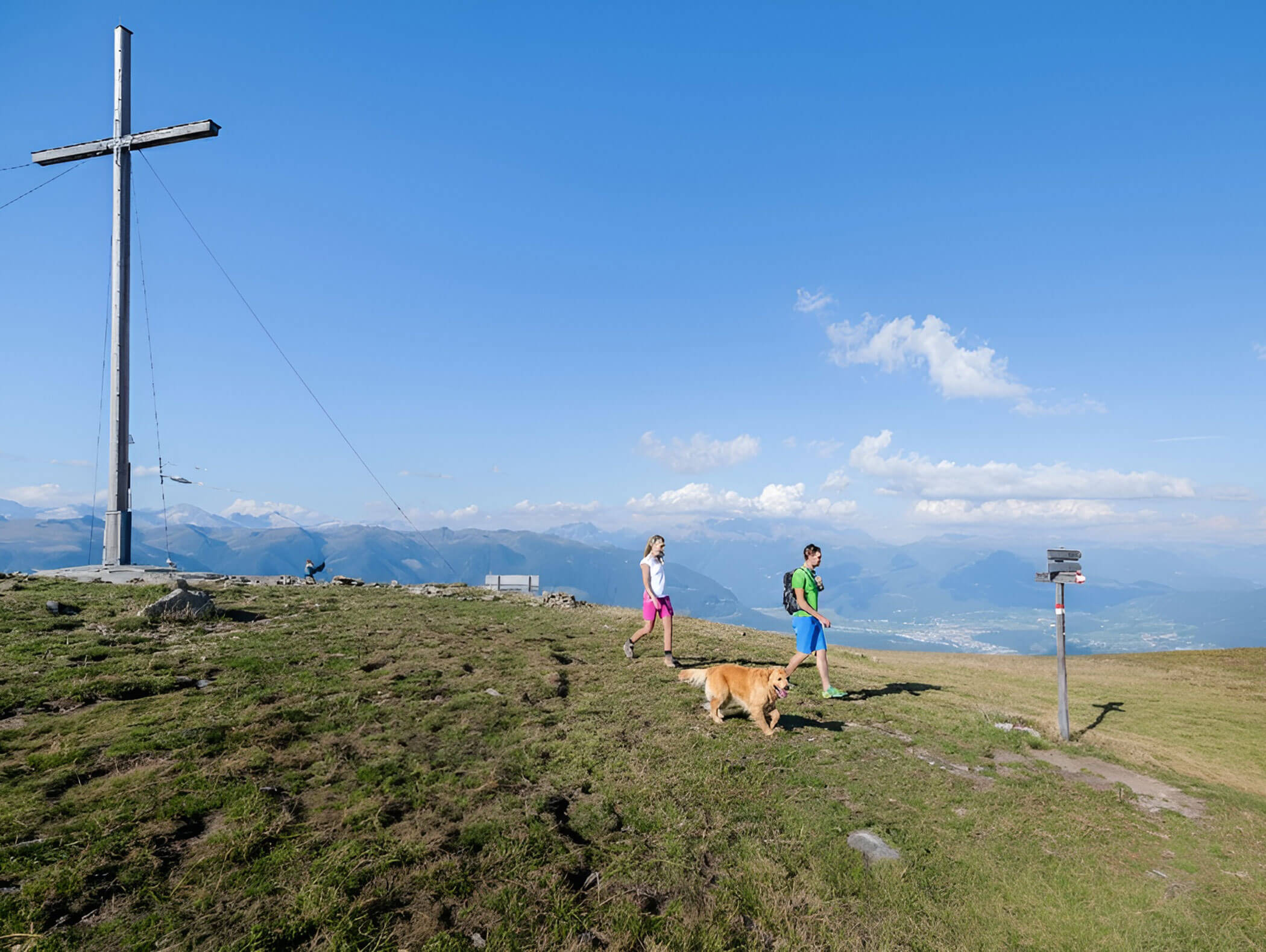 Zwei Menschen und ein Hund spazieren auf einer grasbewachsenen Anhöhe in der Nähe eines großen Kreuzes, mit Bergen und blauem Himmel im Hintergrund. - Hotel B&B MyLiving
