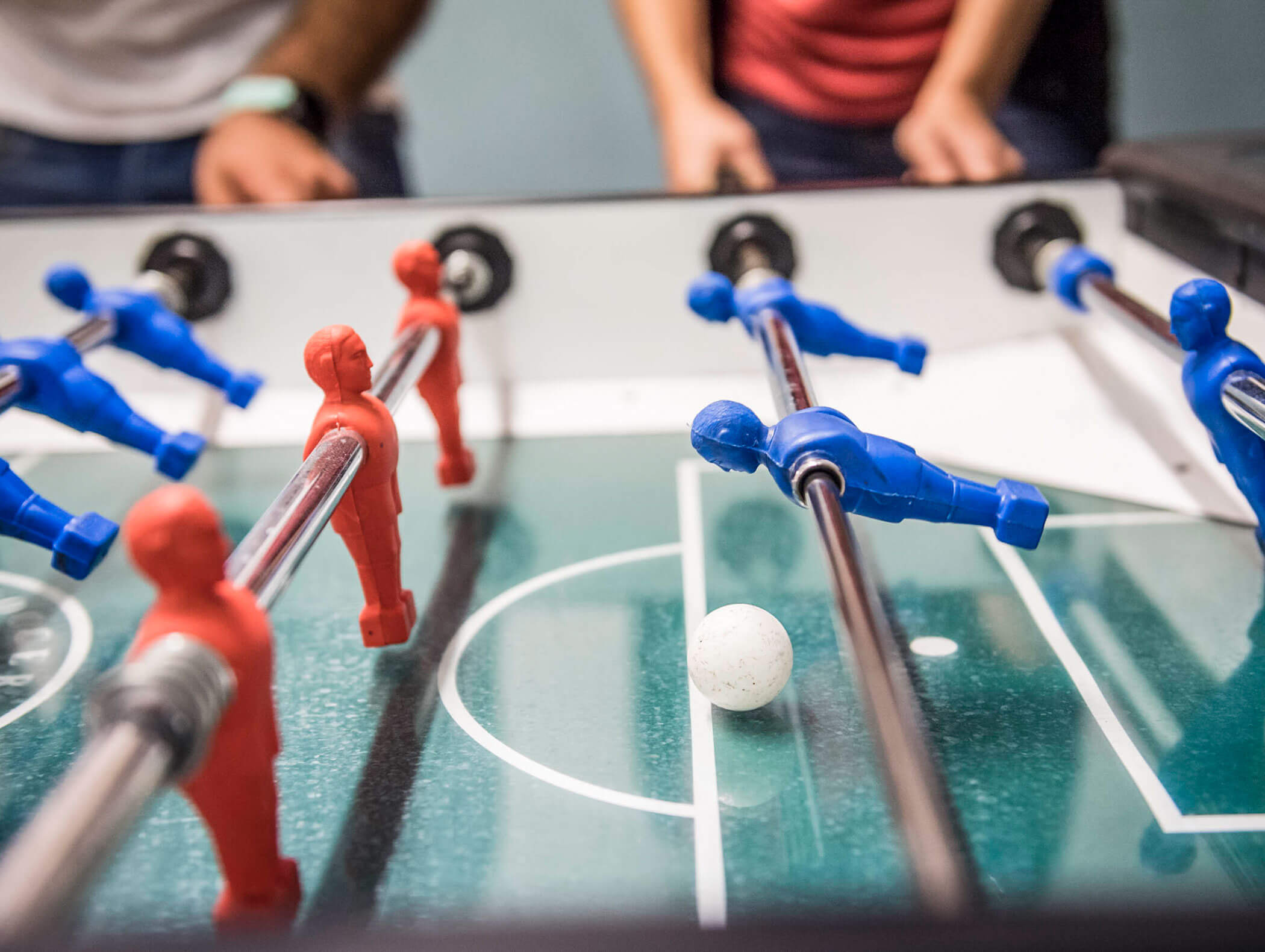 Nahaufnahme eines Kickertisches mit roten und blauen Spielern und einem weißen Ball, im Hintergrund die Hände von Menschen. - Hotel B&B MyLiving