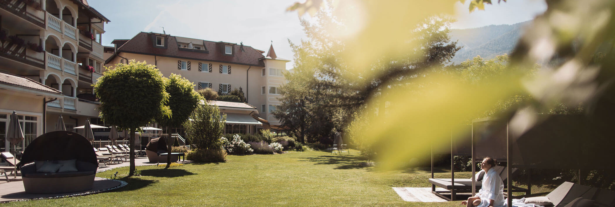 Eine Person in einem weißen Bademantel entspannt sich auf einer Wiese in der Nähe eines Hotels, umgeben von Grün und sonnenbeschienenen Bäumen. - Hotel B&B MyLiving