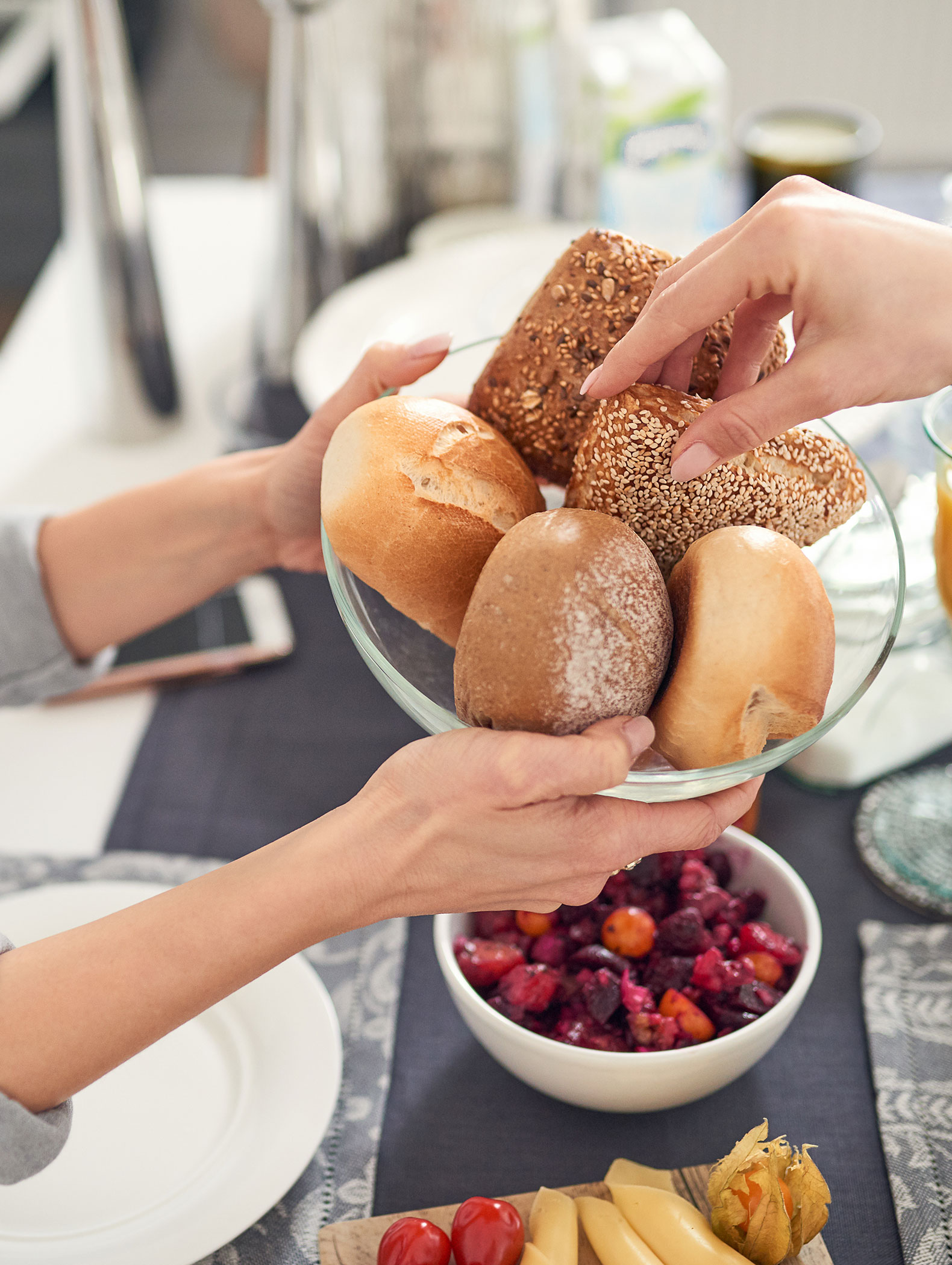 Two hands pass a glass bowl of assorted bread rolls over a table set with food and dishes. - Hotel B&B MyLiving