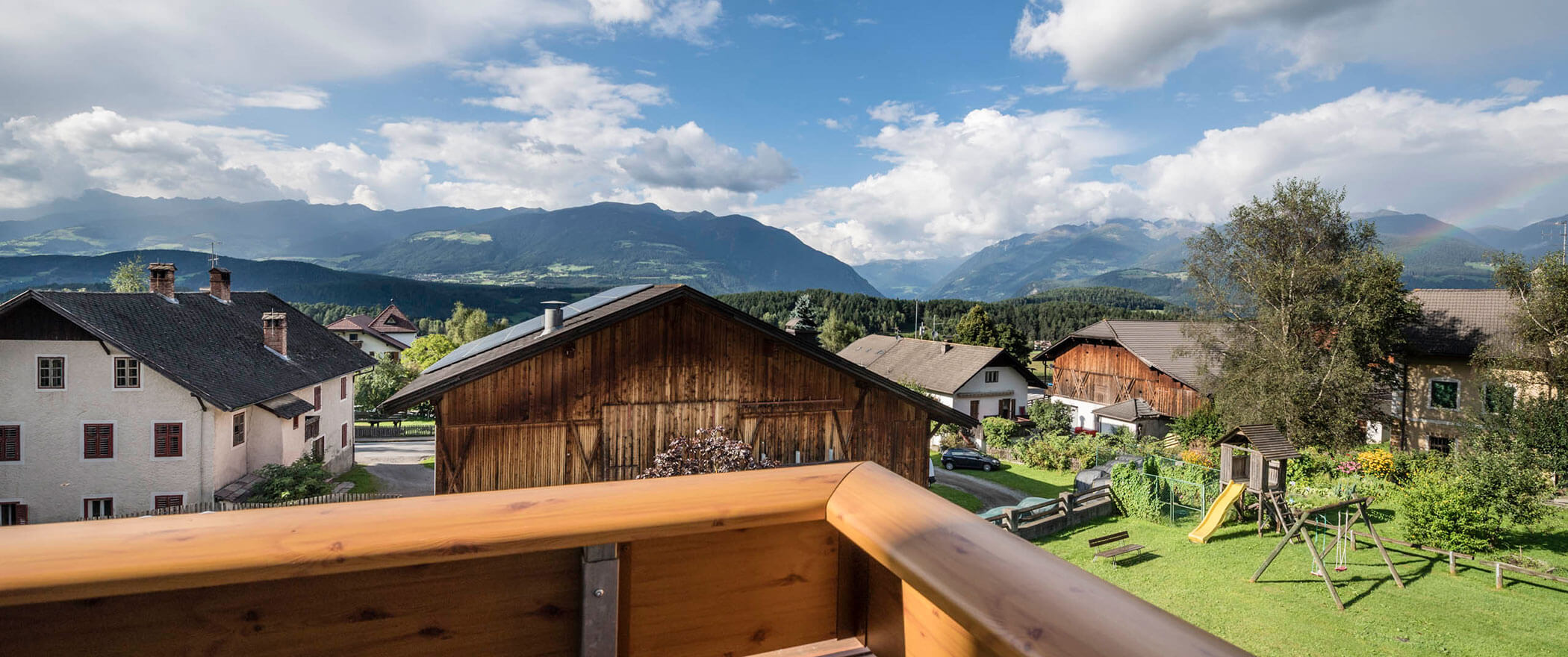 Blick von einem Holzbalkon auf Dorfhäuser, grüne Rasenflächen und ferne Berge bei teilweise bewölktem Himmel. - Hotel B&B MyLiving