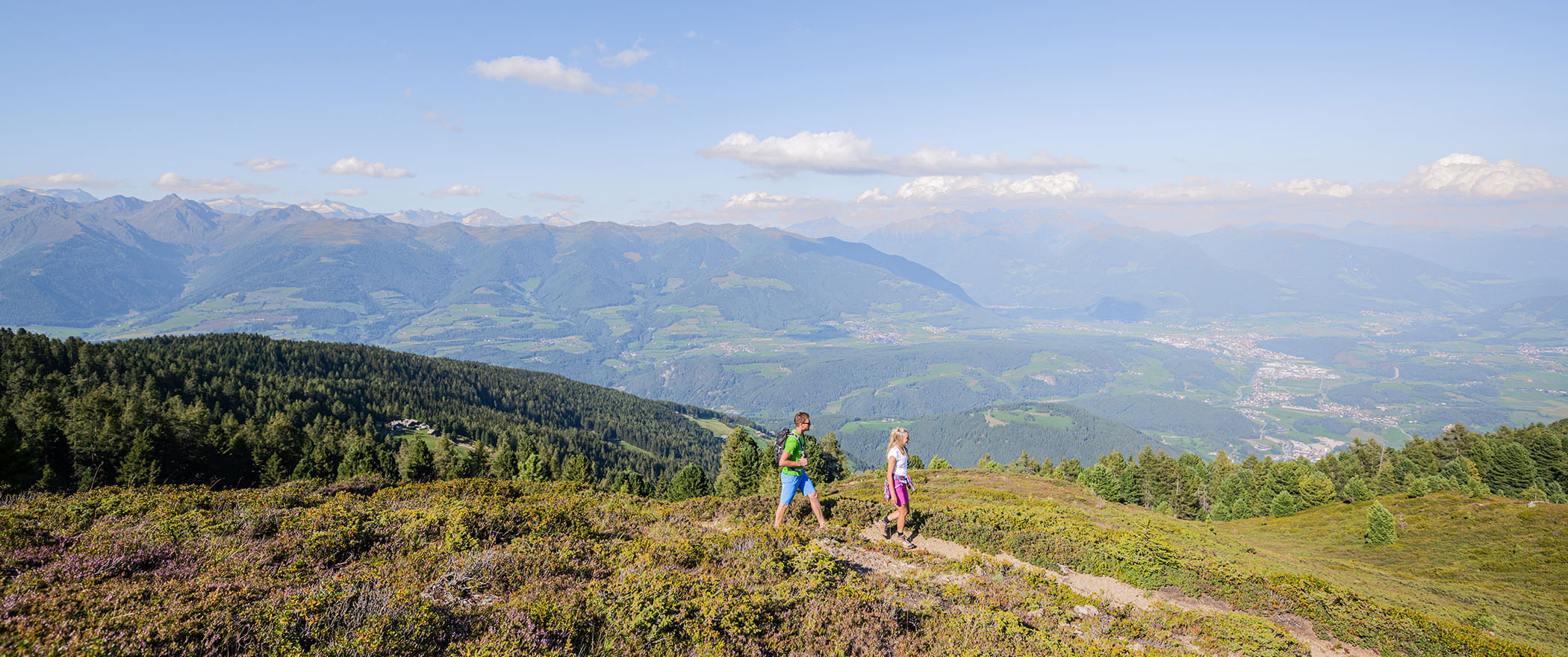 Zwei Personen wandern auf einem malerischen Bergpfad mit grünen Hügeln und einem Tal im Hintergrund. - Hotel B&B MyLiving