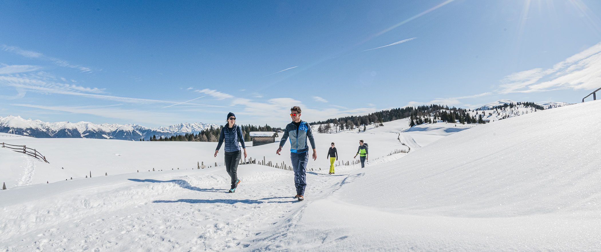 Vier Personen wandern auf einem verschneiten Bergpfad unter einem klaren blauen Himmel mit schneebedeckten Gipfeln im Hintergrund. - Hotel B&B MyLiving
