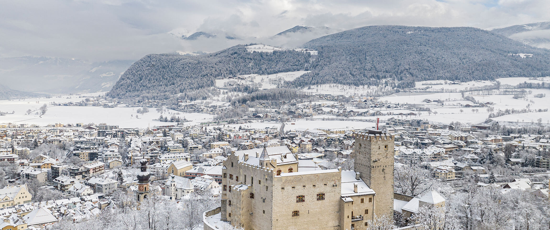Verschneites Schloss und Stadt, umgeben von verschneiten Bergen unter einem bewölkten Winterhimmel. - Hotel B&B MyLiving
