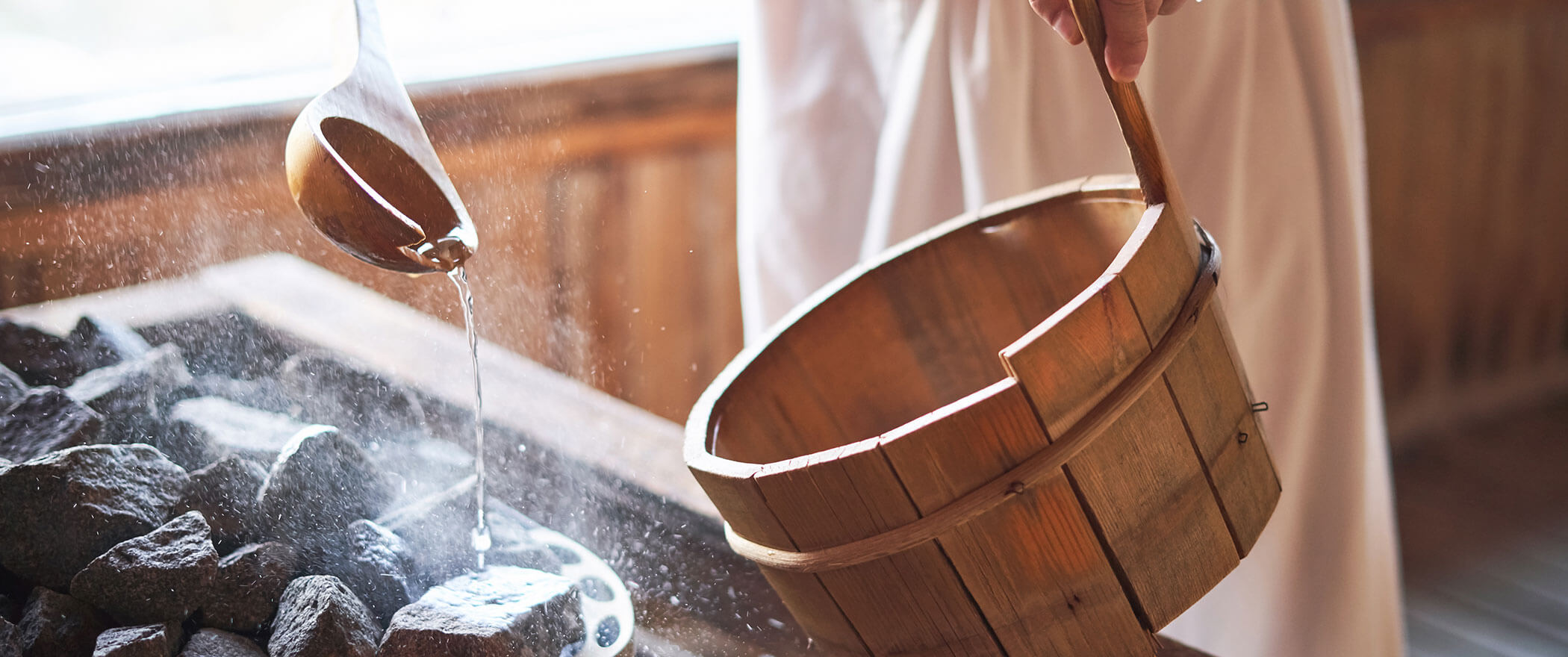 Eine Hand gießt Wasser aus einer Schöpfkelle in einen Holzeimer neben heißen Steinen in einer Sauna. - Hotel B&B MyLiving